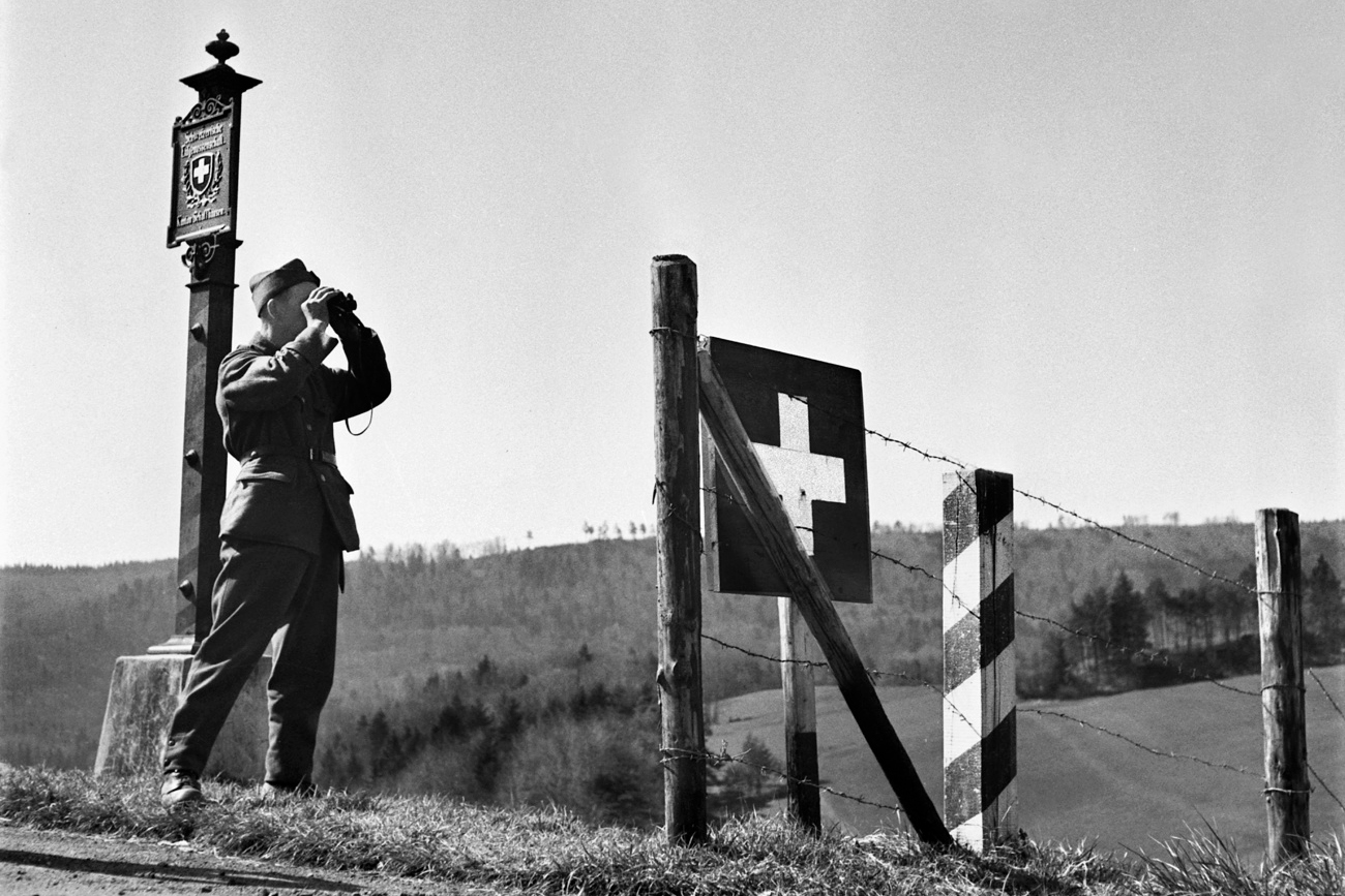 Un soldat se tient devant une clôture en bois à la frontière, marquée d'un drapeau suisse et d'un panneau. Il regarde à travers des jumelles.